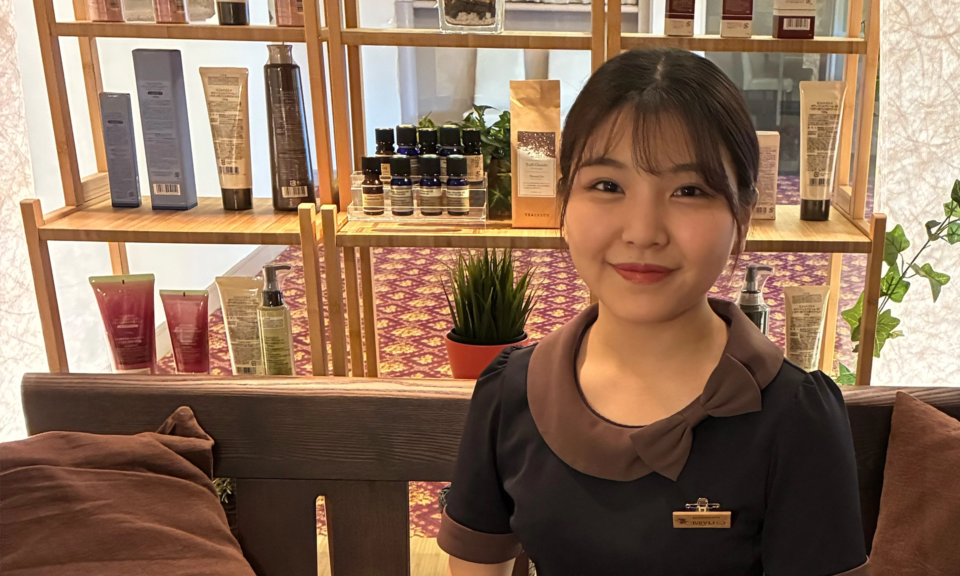 Smiling female spa professional in a uniform, with spa products on shelves in the background, representing a welcoming and professional work environment.
